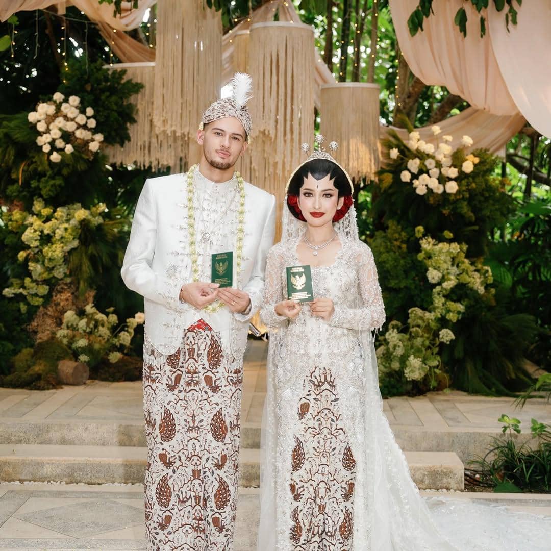 Sepasang pengantin mengenakan busana adat Jawa berwarna putih dan batik cokelat sambil memegang buku nikah di pelaminan berhias bunga.
