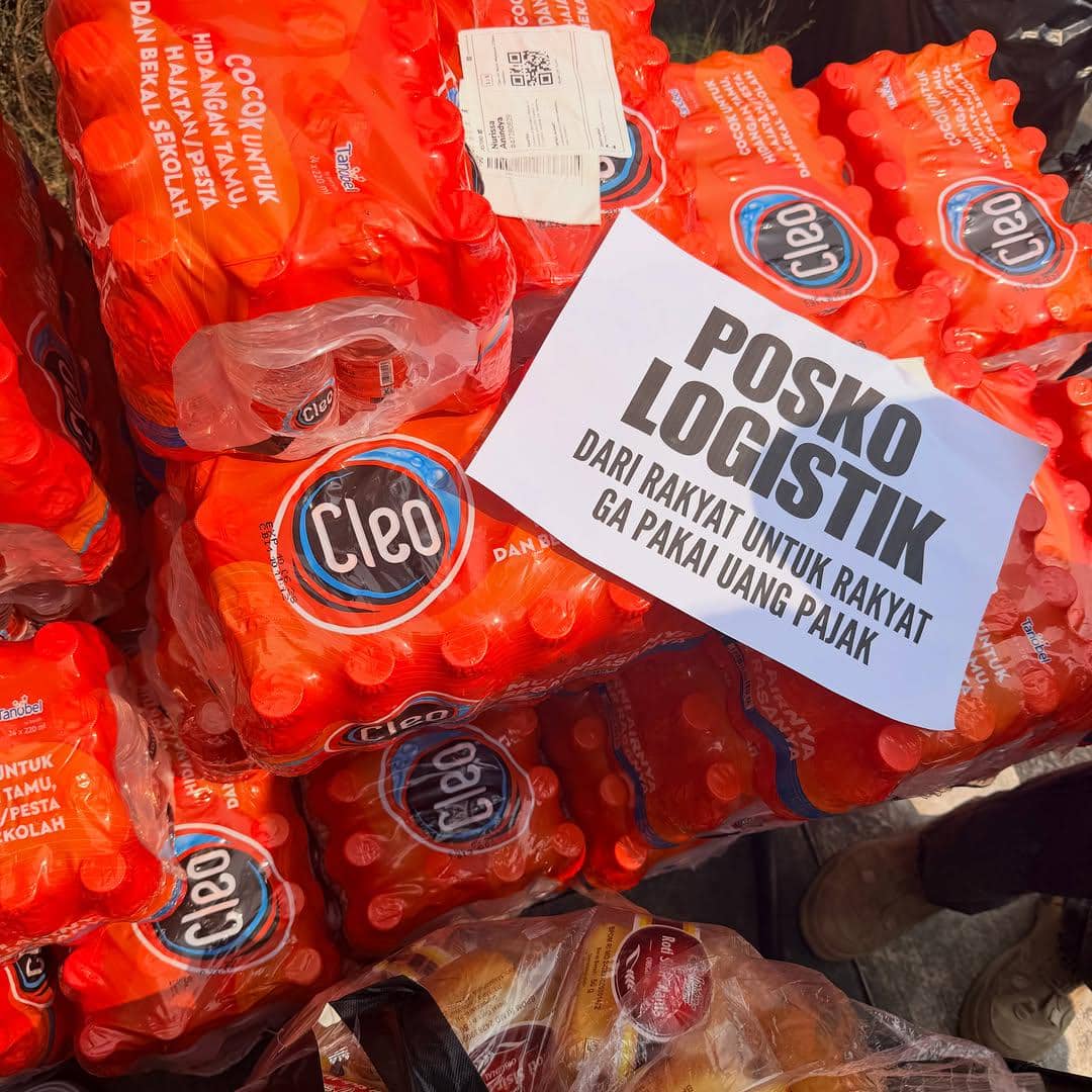 Tumpukan air mineral kemasan Cleo dengan kertas bertuliskan posko logistik dari rakyat untuk rakyat tanpa uang pajak di area demonstrasi.