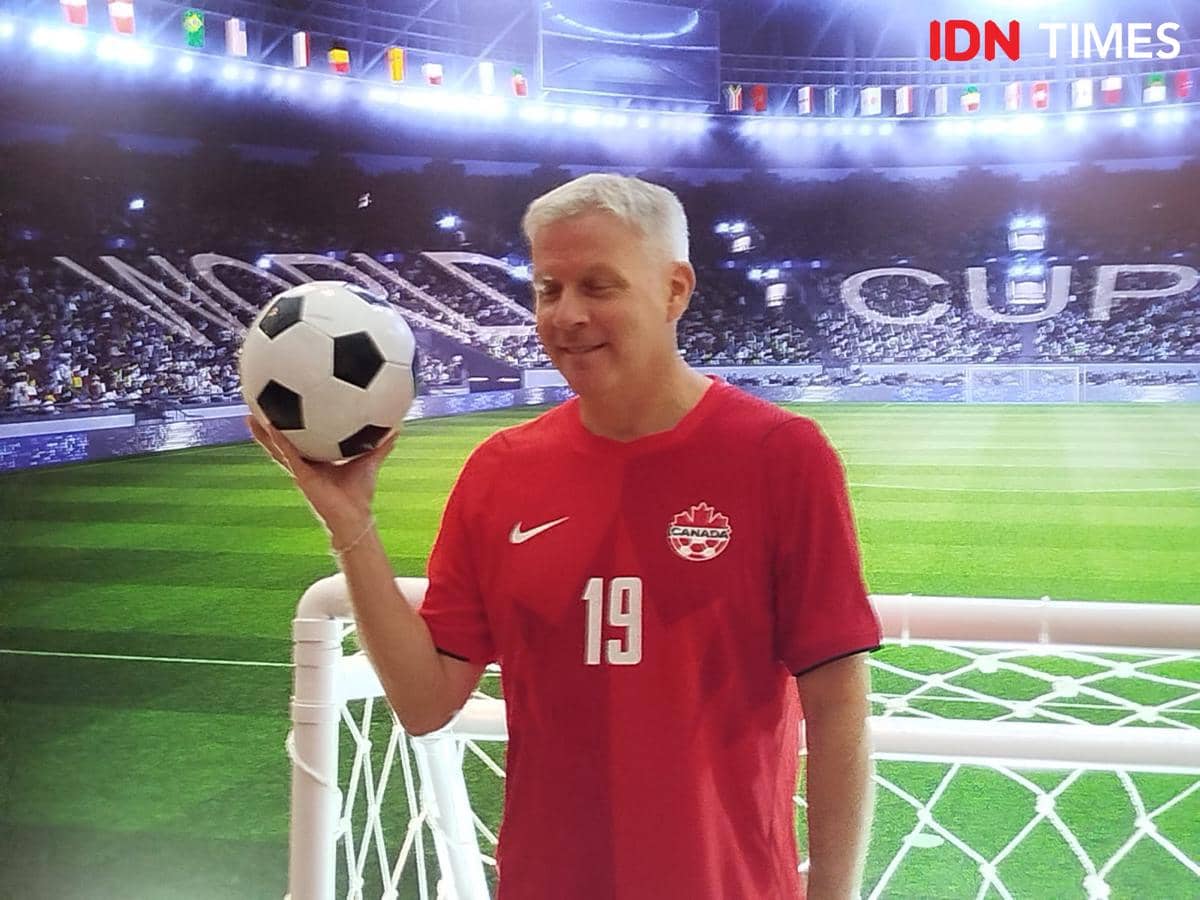 Seorang pria mengenakan jersey tim nasional Kanada berwarna merah memegang bola sepak di depan gawang di stadion bertuliskan World Cup.