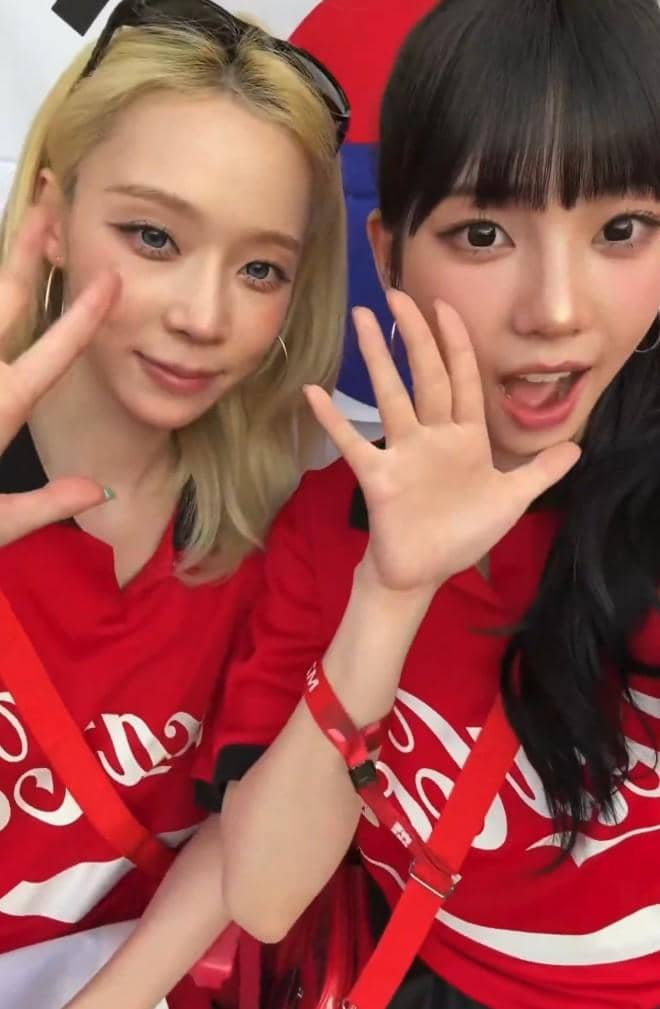 Dua perempuan mengenakan kaus merah dengan latar bendera Korea Selatan, tersenyum dan melambaikan tangan ke kamera.