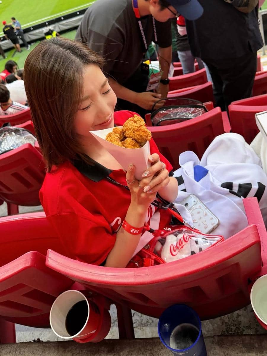 Kwon Eun mengenakan kaus merah duduk di tribun stadion sambil memegang ayam goreng dalam wadah kertas saat menonton pertandingan sepak bola.
