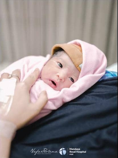 Bayi baru lahir dibedong selimut pink dan mengenakan topi cokelat, terbaring di atas kain biru dengan tangan orang dewasa menyentuhnya.