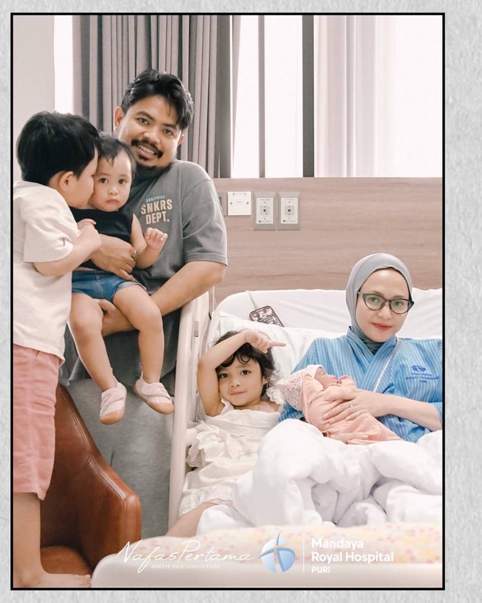 Seorang ibu mengenakan hijab duduk di ranjang rumah sakit sambil menggendong bayi, didampingi suami dan tiga anak mereka di kamar rumah sakit.