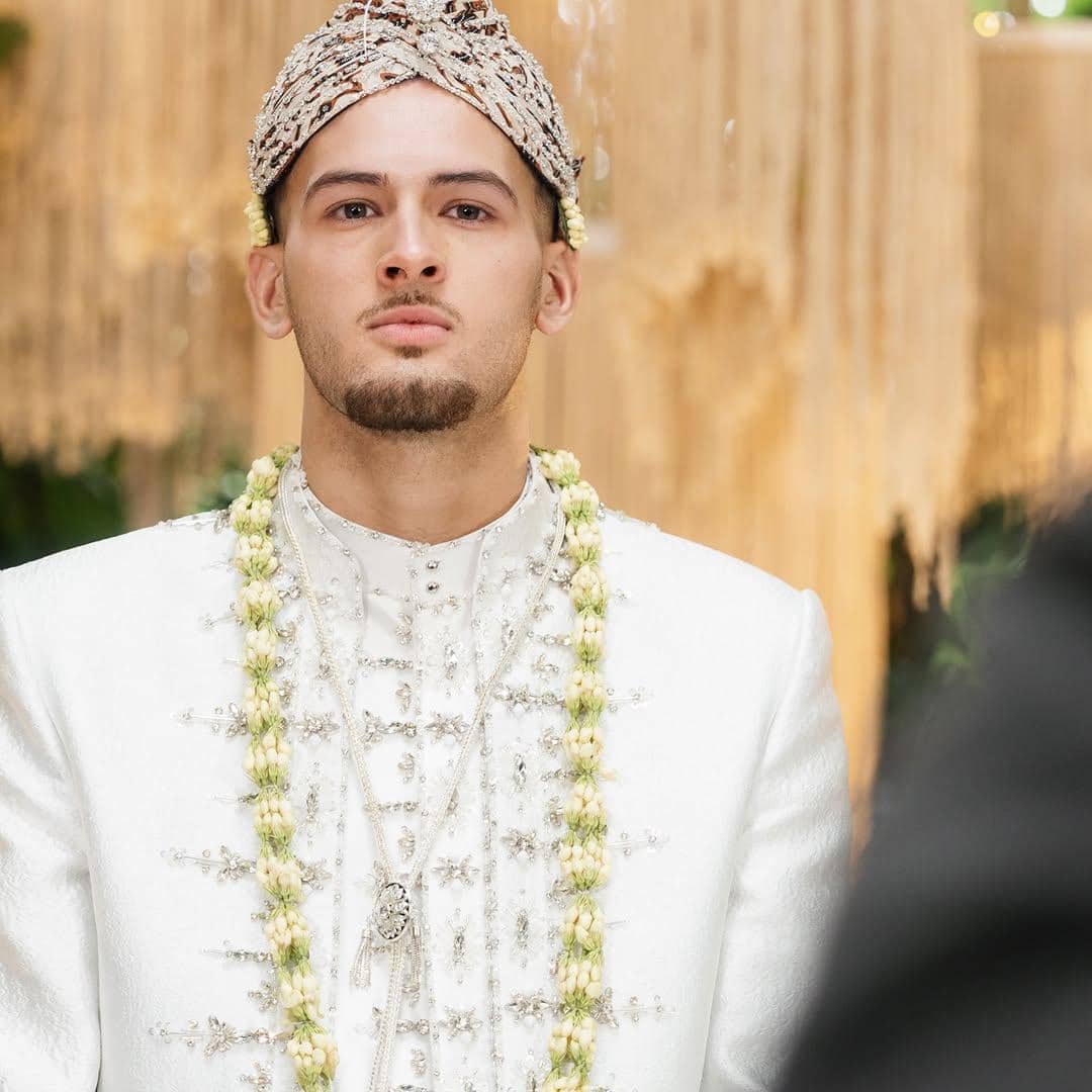 Seorang pria mengenakan busana pengantin adat berwarna putih dengan hiasan melati dan penutup kepala berhias di acara pernikahan.
