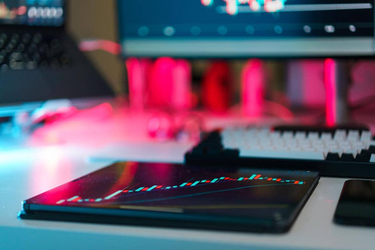 Grafik candlestick berwarna di layar tablet di atas meja kerja dengan pencahayaan neon biru dan merah di latar belakang.