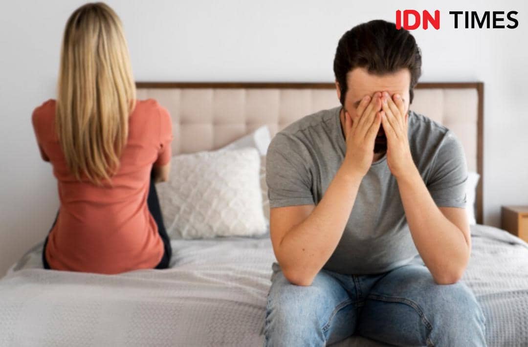 Seorang pria menutupi wajahnya dengan tangan sementara seorang wanita duduk membelakanginya di atas tempat tidur, menggambarkan suasana tegang dalam hubungan.