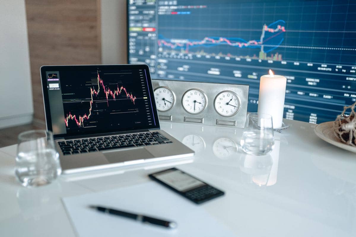 Laptop menampilkan grafik perdagangan saham di meja kerja dengan layar monitor besar, jam dunia, lilin, dan gelas air di sekitarnya.