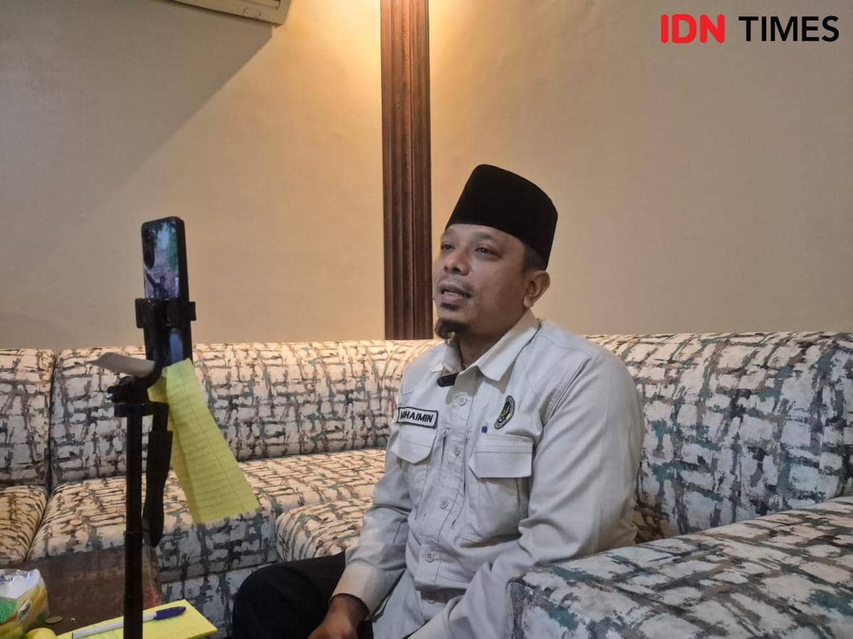 Seorang pria mengenakan kemeja abu-abu dan peci hitam sedang melakukan panggilan video menggunakan ponsel di ruang tamu.