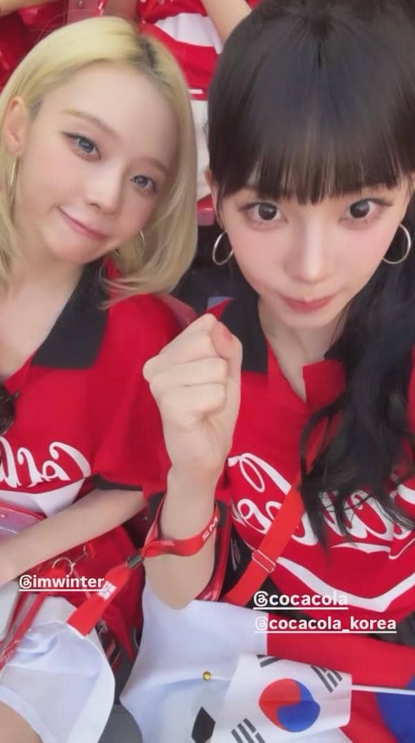 Dua perempuan mengenakan jersey merah dengan logo Coca-Cola dan membawa bendera Korea Selatan saat menonton pertandingan sepak bola.