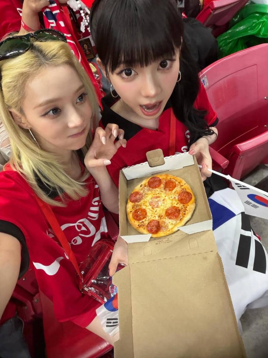 Winter dan Karina aespa mengenakan kaus merah menonton pertandingan sepak bola sambil memegang kotak pizza kecil di tribun stadion.