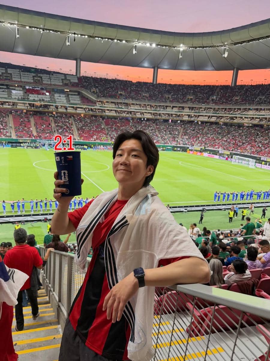 Seunghoon WINNER mengenakan kaus merah dan syal putih berpose di stadion saat pertandingan sepak bola Korea Selatan di Meksiko.