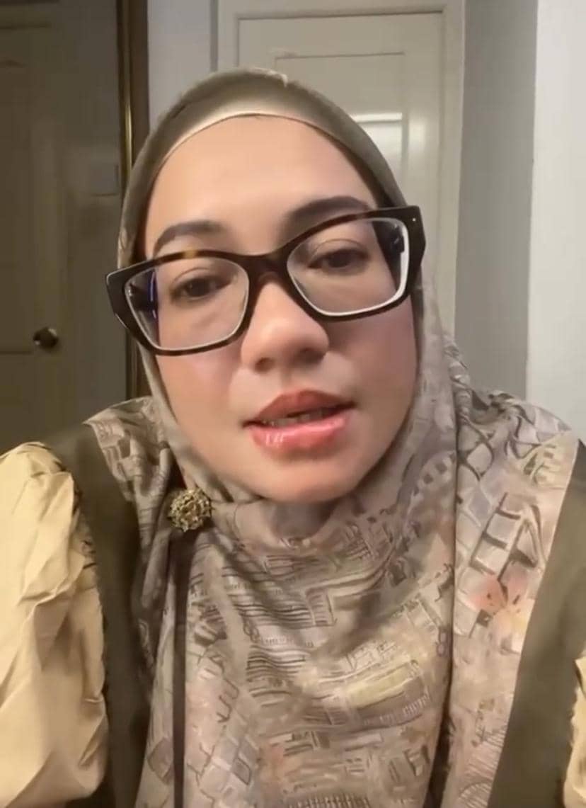 Seorang perempuan berhijab bermotif dan berkacamata besar sedang menatap kamera di dalam ruangan dengan pencahayaan hangat.