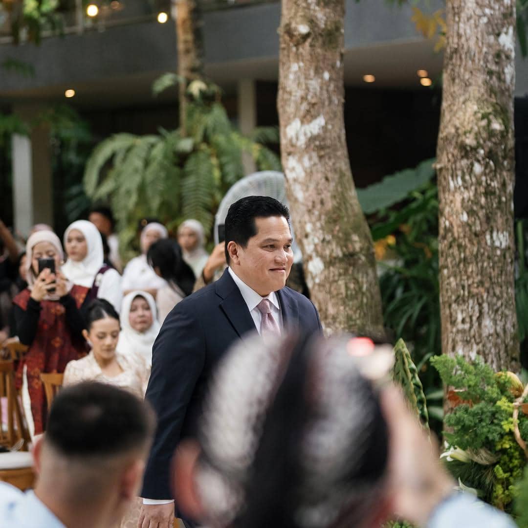 Seorang pria mengenakan setelan jas gelap dan dasi berjalan di acara akad nikah dengan tamu undangan di latar belakang.