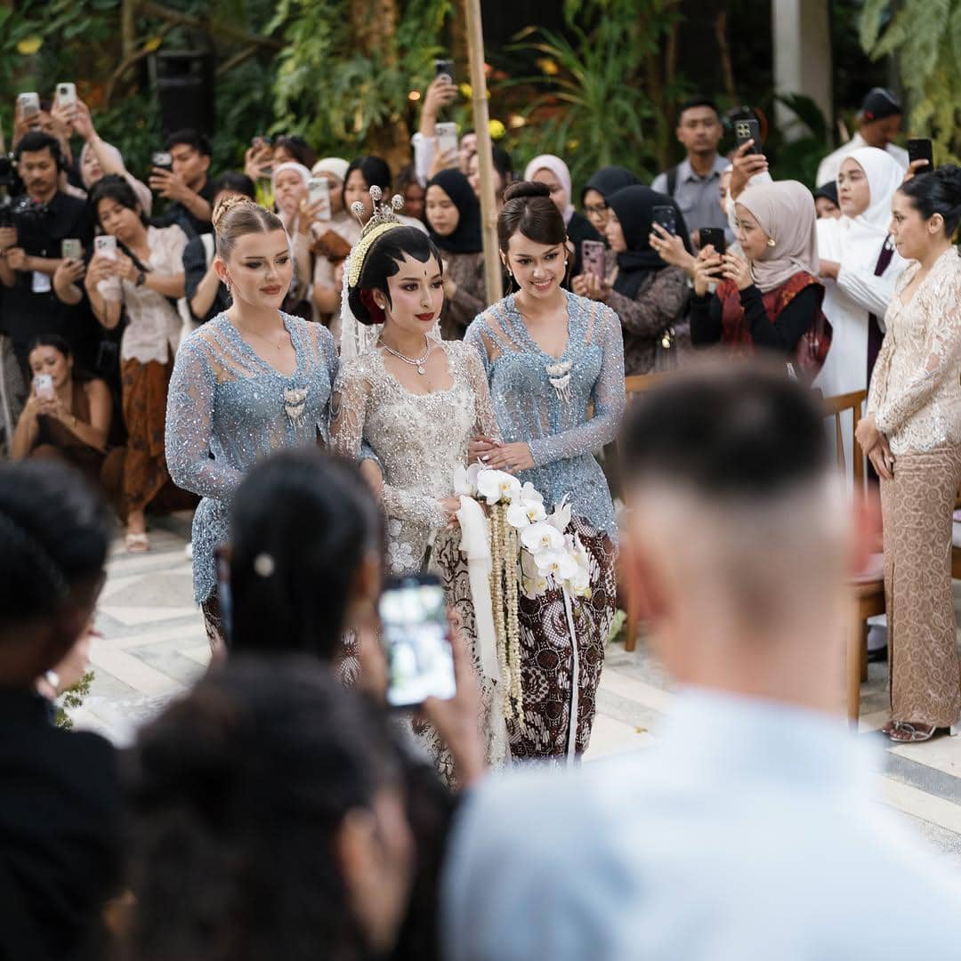 Seorang pengantin wanita mengenakan kebaya putih dan siger berjalan diiringi dua perempuan berkebaya biru dalam prosesi akad nikah di depan tamu undangan.
