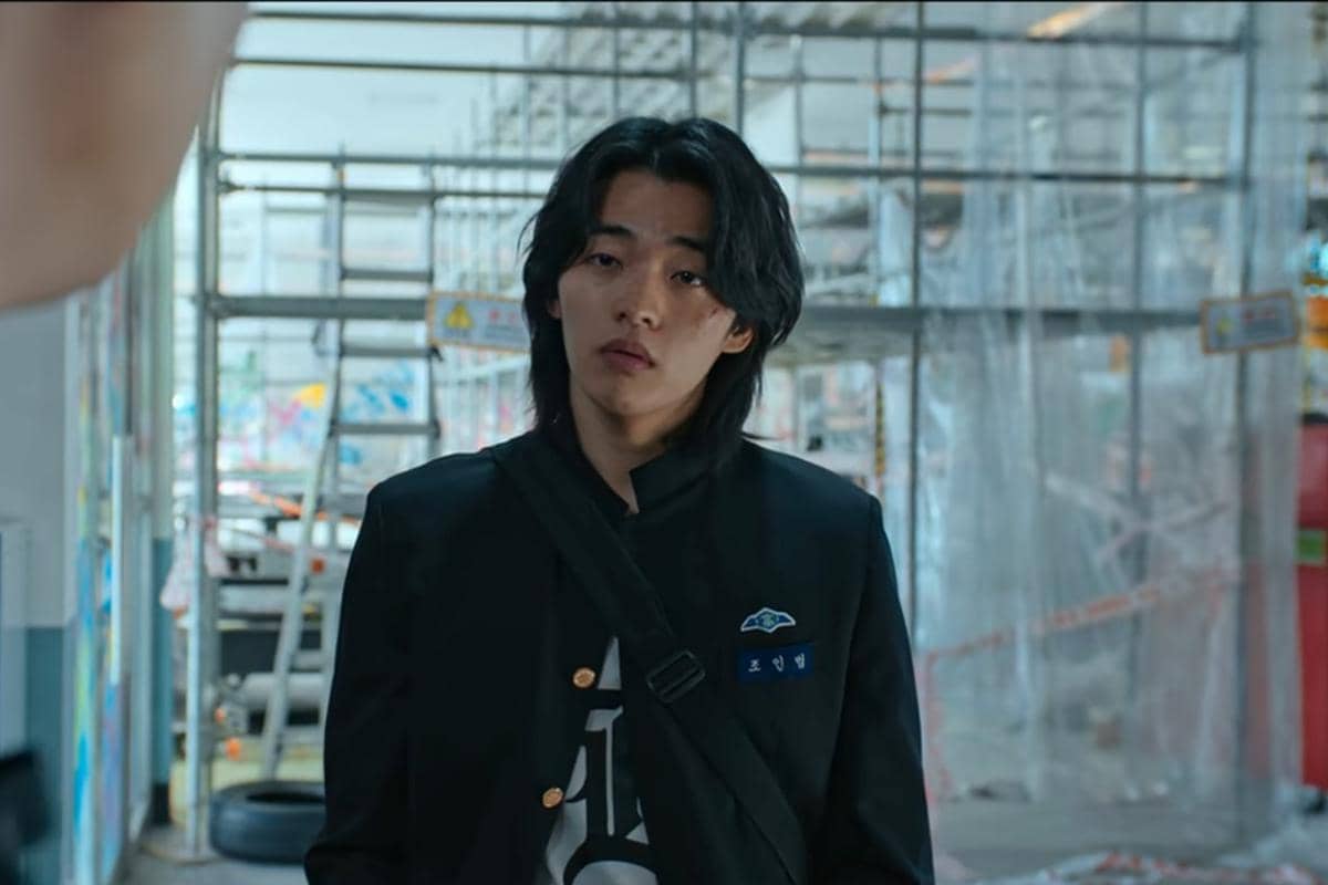 Seorang siswa mengenakan seragam sekolah hitam dengan tas selempang berdiri di area bangunan dengan perancah di drama Korea Teach You a Lesson.
