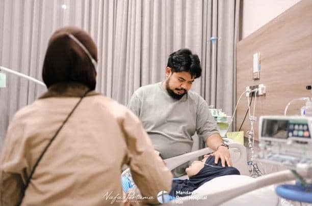 Seorang pria duduk di samping tempat tidur rumah sakit sambil menggenggam tangan seorang wanita yang terbaring, dengan suasana hangat dan penuh perhatian.