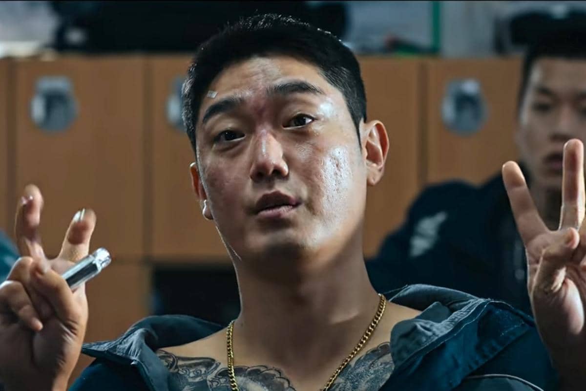 Seorang pria berambut pendek dengan tato di dada dan kalung emas mengangkat kedua tangannya membentuk tanda kutip di adegan drama Korea Teach You a Lesson.