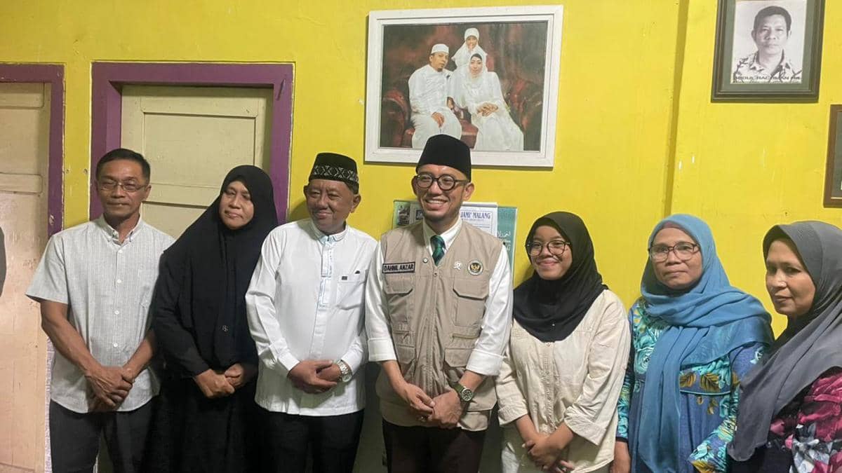 Seorang pejabat mengenakan rompi resmi berfoto bersama keluarga di dalam rumah dengan dinding kuning dan potret keluarga di belakangnya.