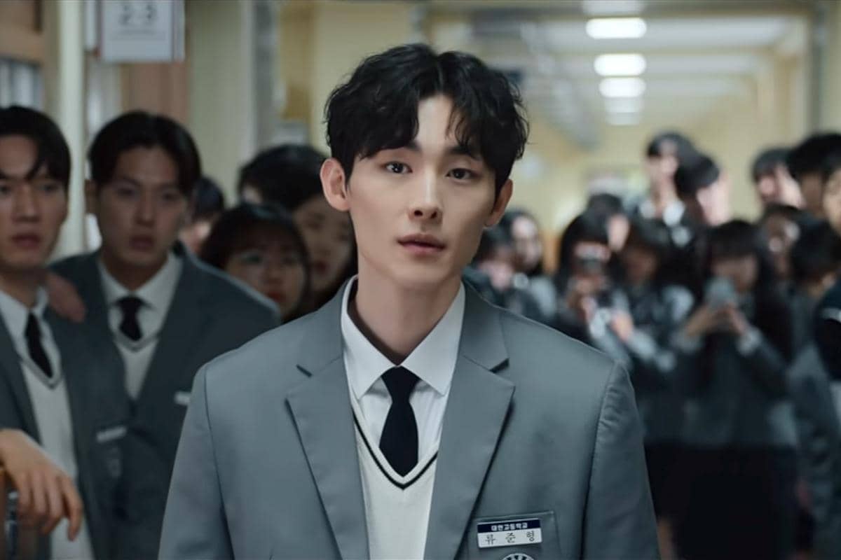 Seorang siswa berseragam abu-abu berjalan di koridor sekolah dengan ekspresi serius, dikelilingi oleh siswa lain dalam adegan drama Korea Teach You a Lesson.