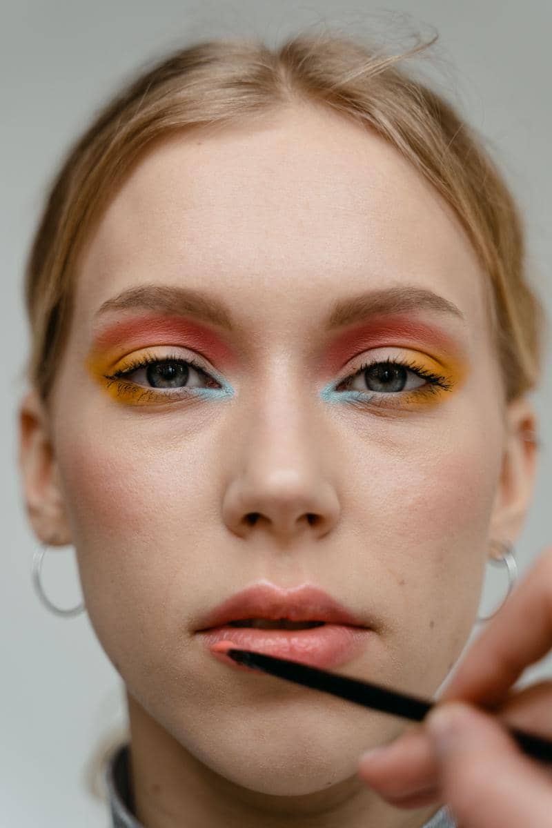Wajah seorang perempuan dengan riasan mata warna-warni bernuansa merah, kuning, dan biru sedang dirias bibirnya dengan kuas.