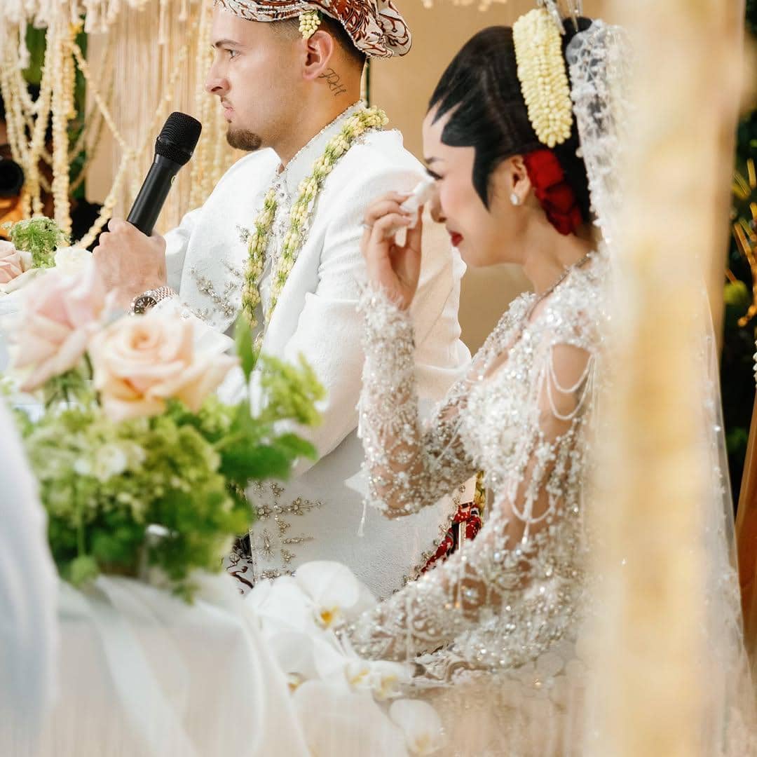 Pasangan pengantin mengenakan busana adat Jawa berwarna putih saat prosesi akad nikah dengan dekorasi bunga segar di sekitarnya.