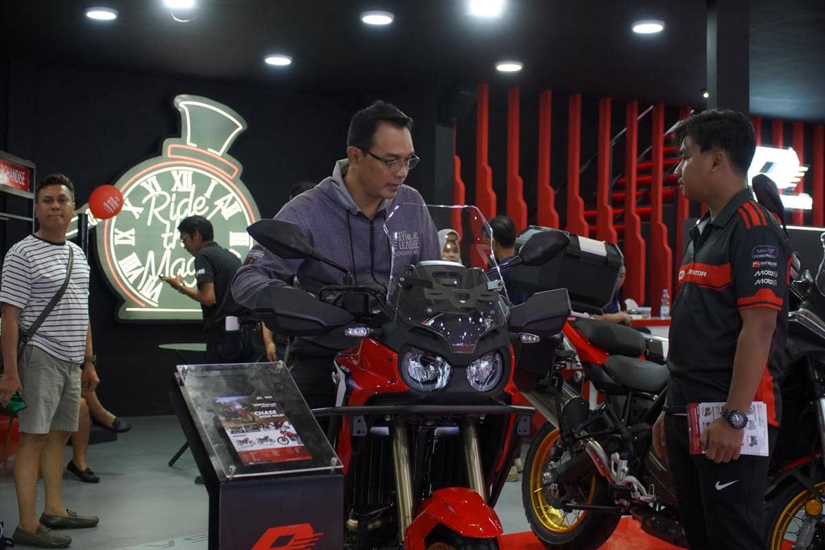 Seorang pengunjung melihat motor di booth QJMotor pada acara JFK 2026 dengan latar desain modern berwarna merah dan hitam.