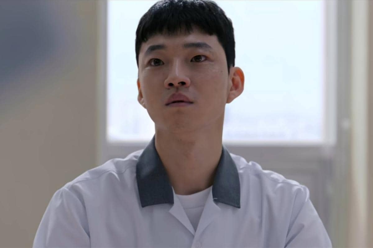Seorang pria mengenakan seragam putih dengan kerah abu-abu duduk di dalam ruangan terang dalam adegan drama Korea Teach You a Lesson.