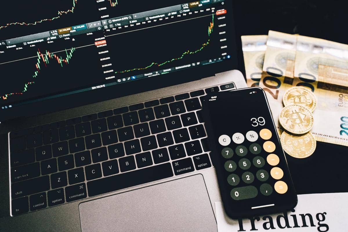 Laptop menampilkan grafik perdagangan dengan ponsel kalkulator di atasnya, disertai uang kertas euro dan koin bitcoin di sampingnya.