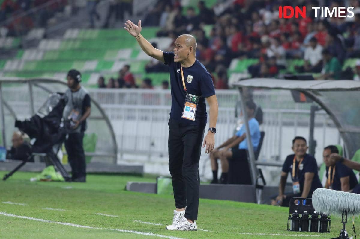 Seorang pelatih sepak bola memberikan instruksi di pinggir lapangan saat pertandingan berlangsung di stadion dengan penonton di tribun.