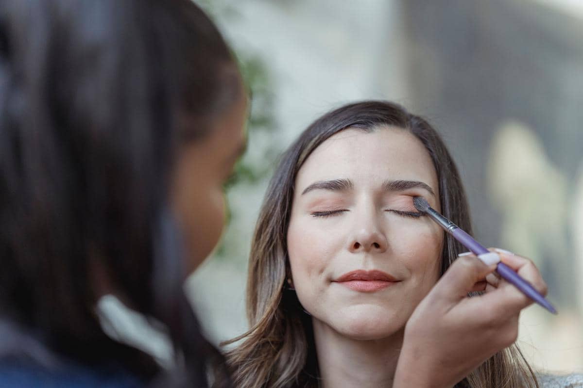 Seorang perempuan dengan mata terpejam sedang dirias oleh penata rias yang mengaplikasikan eyeshadow menggunakan kuas makeup.