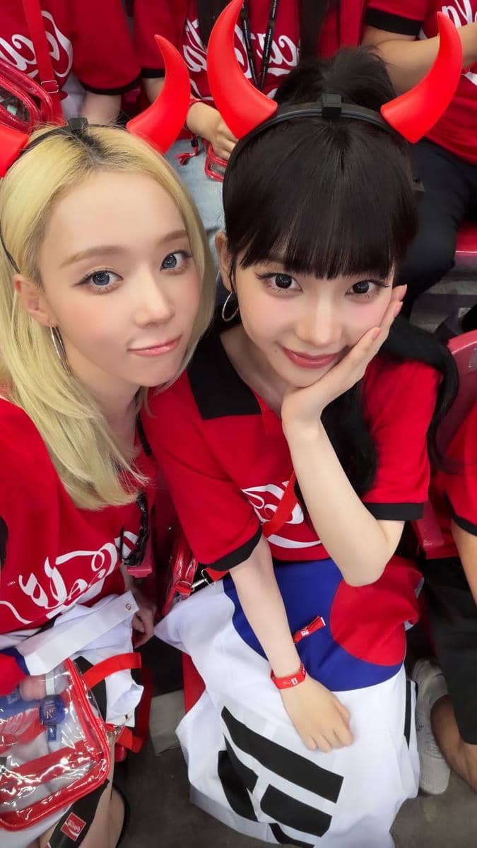 Dua perempuan mengenakan kaus merah dan ikat kepala bertanduk merah berpose di tribun stadion saat mendukung Korea Selatan di Piala Dunia 2026.