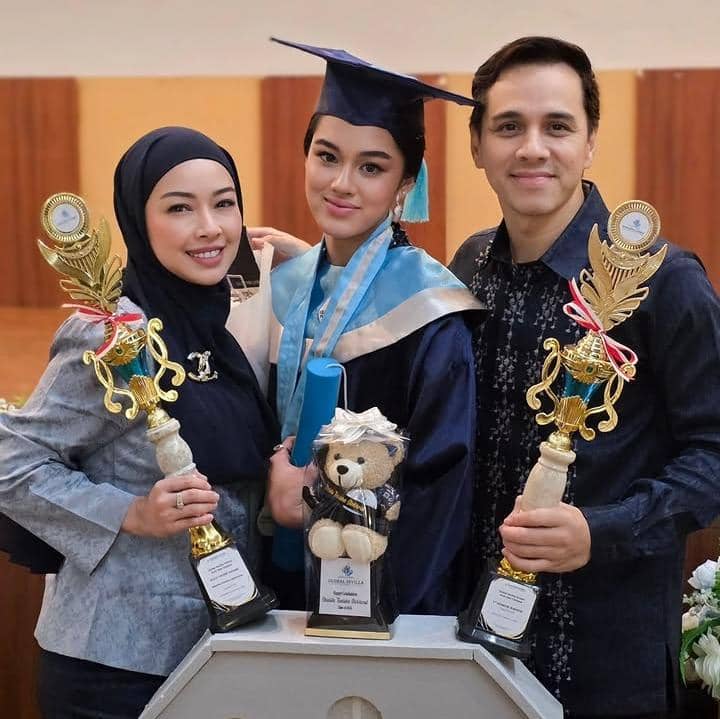 Seorang siswi SMA mengenakan toga wisuda berpose bersama dua orang dewasa yang memegang piala penghargaan di acara kelulusan.