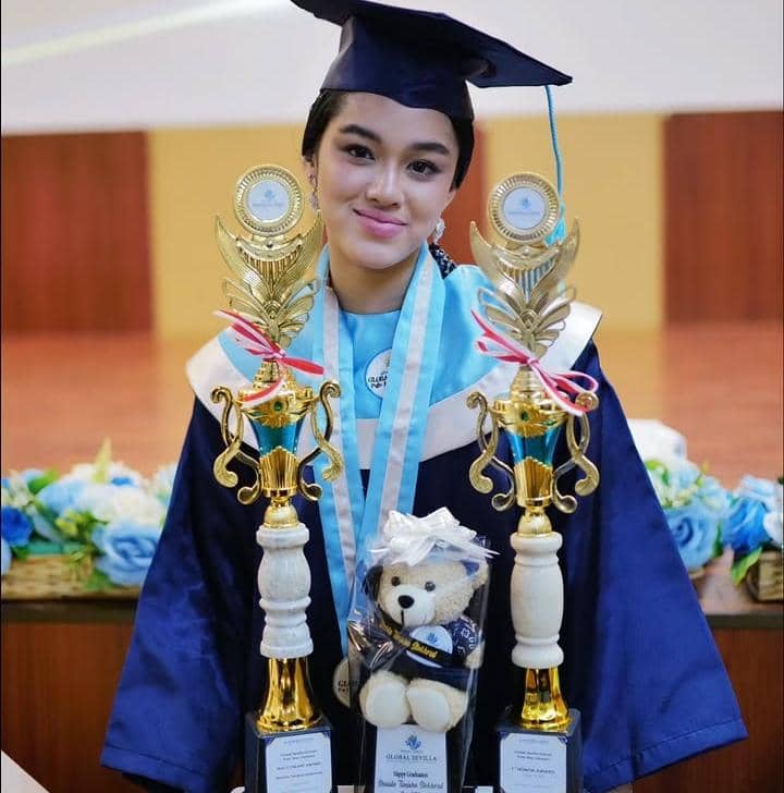 Seorang siswi mengenakan toga wisuda SMA berwarna biru muda dan biru tua sambil memegang dua piala penghargaan di aula sekolah.