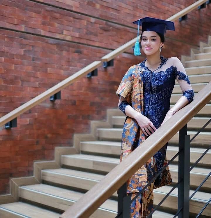 Seorang siswi mengenakan kebaya biru dan kain batik dengan toga wisuda berdiri di tangga dengan latar dinding bata merah.