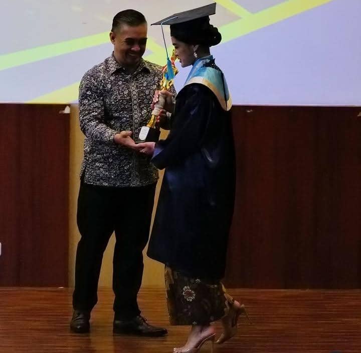 Seorang siswi SMA mengenakan toga wisuda menerima piala dari seorang pria di atas panggung dalam acara kelulusan sekolah.