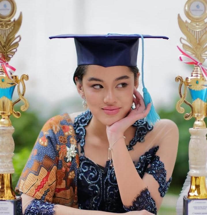 Seorang siswi mengenakan toga wisuda dan kebaya biru dengan selendang batik, duduk di antara dua piala penghargaan.