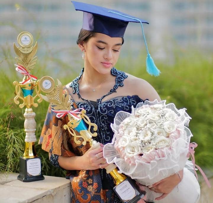 Seorang siswi mengenakan toga wisuda dan kebaya biru memegang buket bunga serta dua piala penghargaan di luar ruangan.