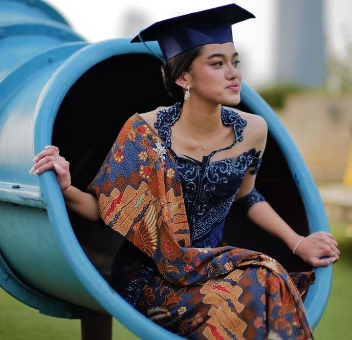 Seorang siswi mengenakan kebaya biru dan selendang batik dengan topi toga duduk di dalam perosotan saat acara wisuda SMA.