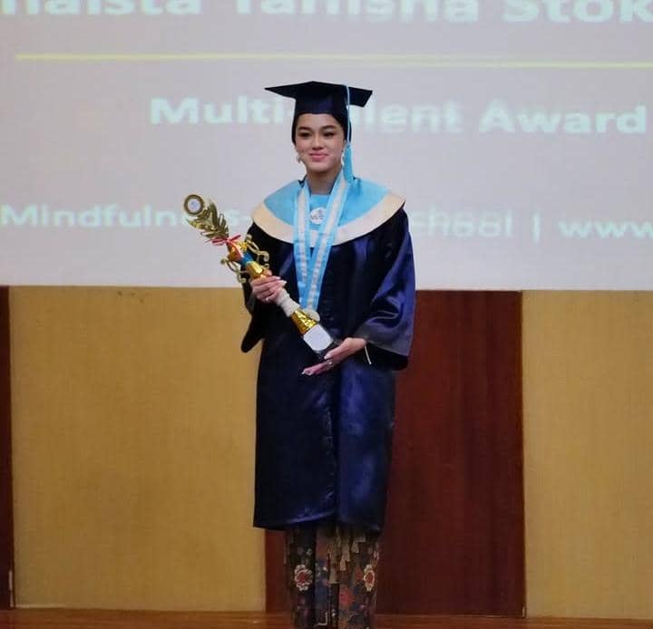 Seorang siswi SMA mengenakan toga wisuda berwarna biru tua sambil memegang piala penghargaan di atas panggung acara kelulusan.
