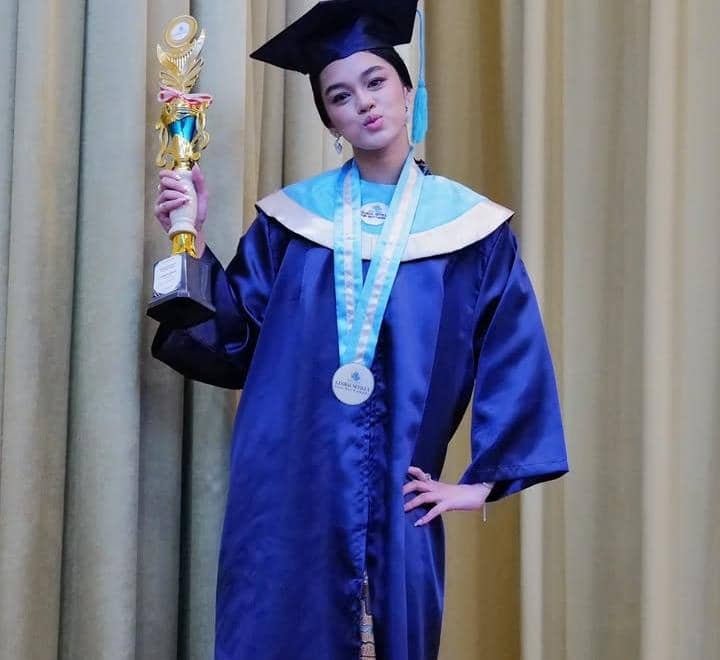 Seorang siswi mengenakan toga wisuda berwarna biru sambil memegang piala dan mengenakan medali di lehernya di depan tirai.