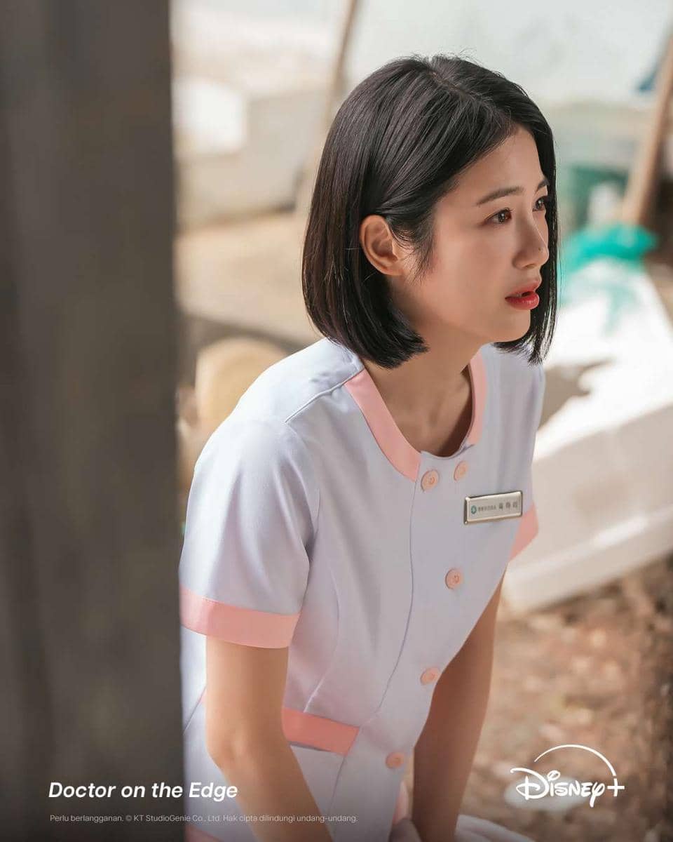 Seorang perempuan berambut pendek mengenakan seragam perawat putih dengan garis merah muda sedang duduk di luar ruangan.