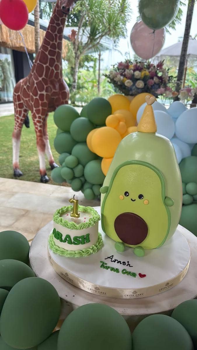 Kue ulang tahun pertama bertema alpukat dengan tulisan Arash di atas meja, dikelilingi balon hijau dan dekorasi pesta anak.