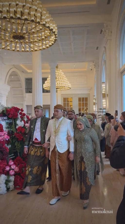 Rombongan pengantin mengenakan busana adat Jawa berjalan di aula megah dengan lampu gantung besar dan dekorasi bunga merah putih.