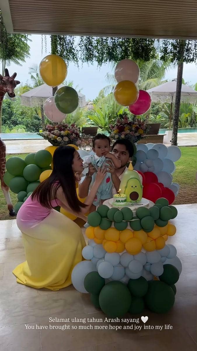 Seorang wanita dan pria merayakan ulang tahun pertama bayi mereka bernama Arash dengan dekorasi balon warna-warni dan kue bertema alpukat.
