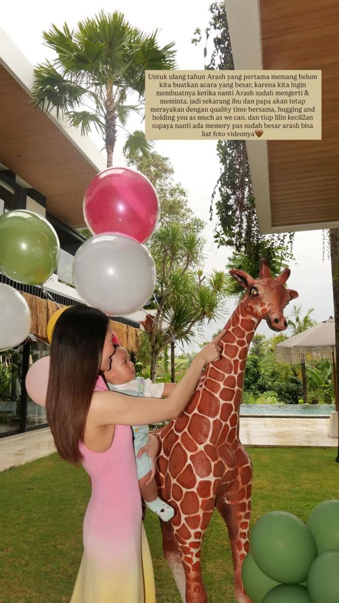 Seorang wanita menggendong bayi sambil memegang patung jerapah di taman dengan balon warna-warni saat perayaan ulang tahun pertama.