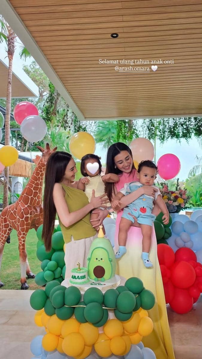 Dua wanita tersenyum sambil menggendong dua anak kecil di pesta ulang tahun pertama bertema alpukat dengan balon warna-warni.