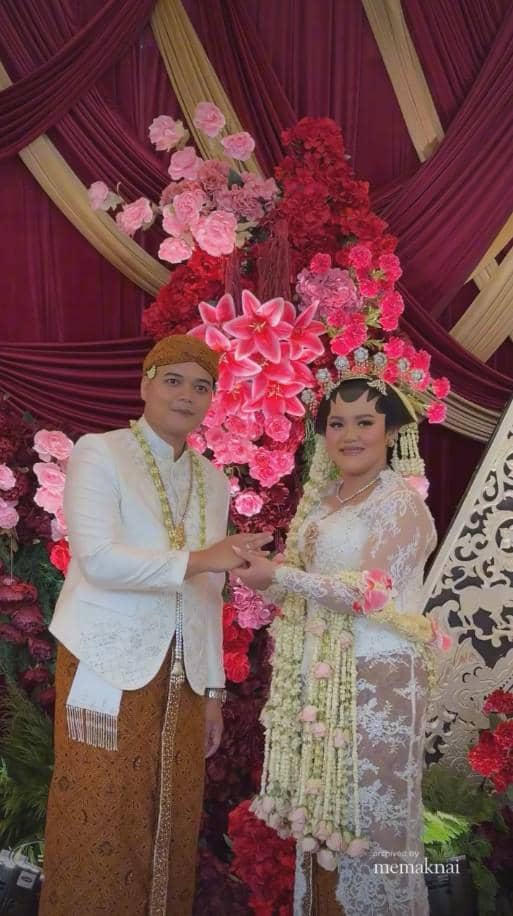 Pasangan pengantin mengenakan busana adat Jawa berwarna putih dan cokelat berpose di pelaminan berhias bunga merah muda dan merah.