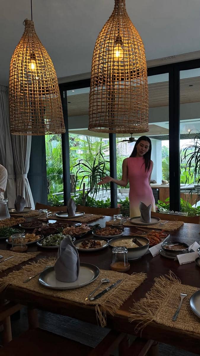 Seorang wanita mengenakan gaun merah muda berdiri di samping meja makan besar yang dipenuhi hidangan di ruangan bernuansa alami dengan lampu rotan gantung.