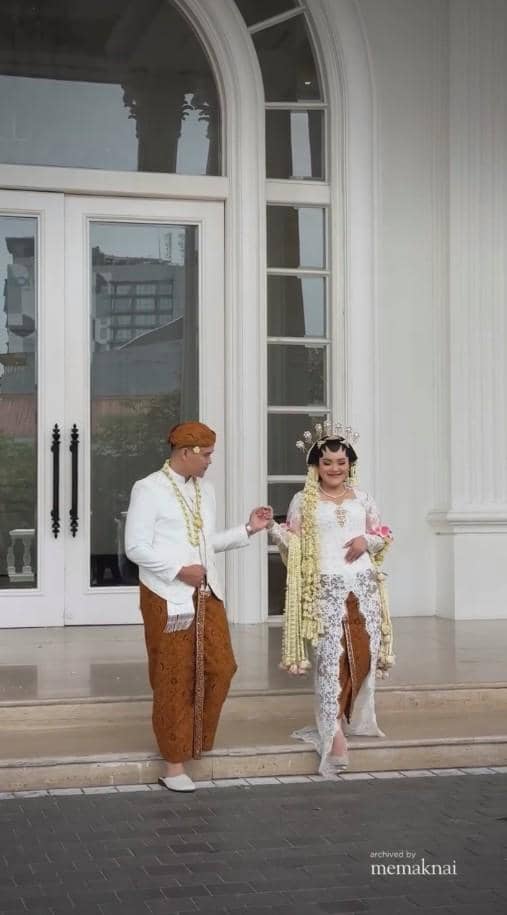 Pasangan pengantin mengenakan busana adat Jawa, pengantin pria bersetelan putih dengan kain batik dan pengantin wanita berkebaya putih berhias ronce melati.