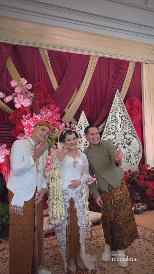 Pasangan pengantin mengenakan busana adat Jawa berpose bersama tamu di pelaminan berhias bunga merah dan dekorasi tradisional.